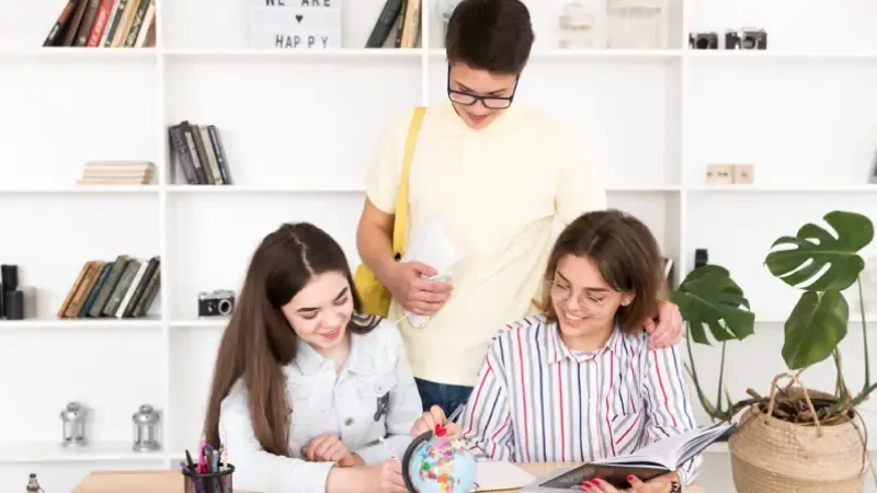 A group of three people engaged in studying, surrounded by books, a globe, and a potted plant in a bright, modern room